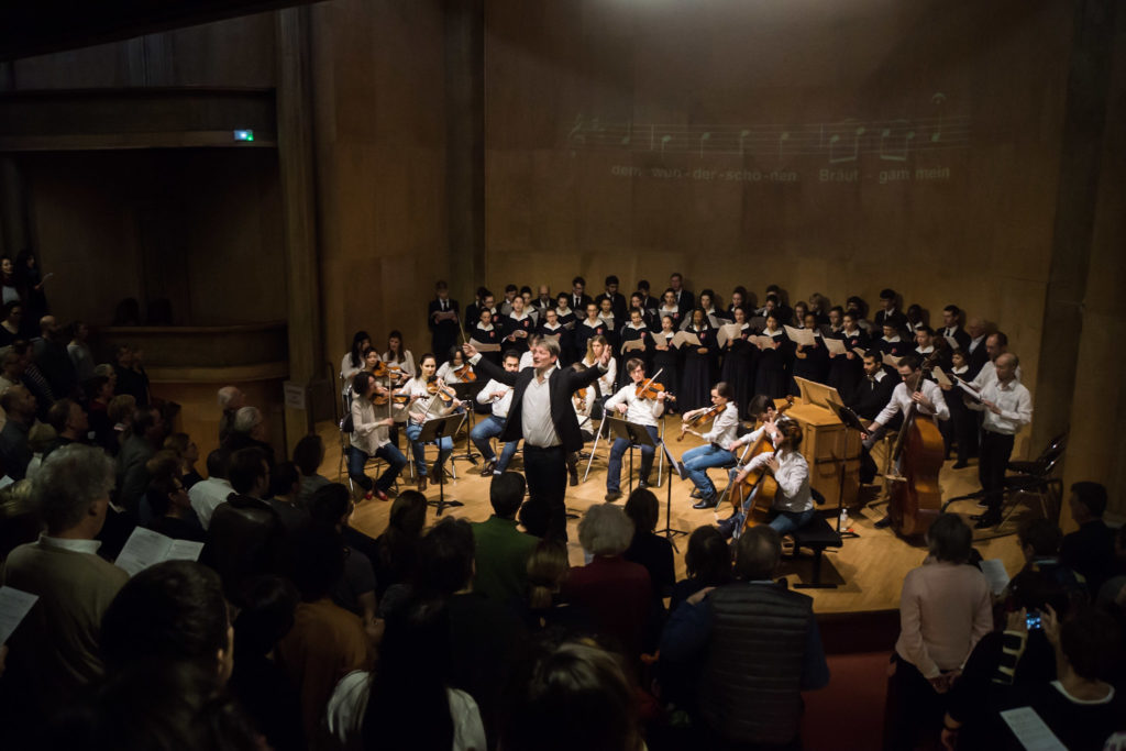 Une représentation du concert Bach and Breakfast avec orchestre baroque chœurs et solistes chanteurs. Le chef d'orchestre Jérôme Pernoo est tourné vers le public et le dirige pour chanter un choral de Jean-Sébastien Bach. Les notes et les paroles s'affichent sur le mur de fond de scène.