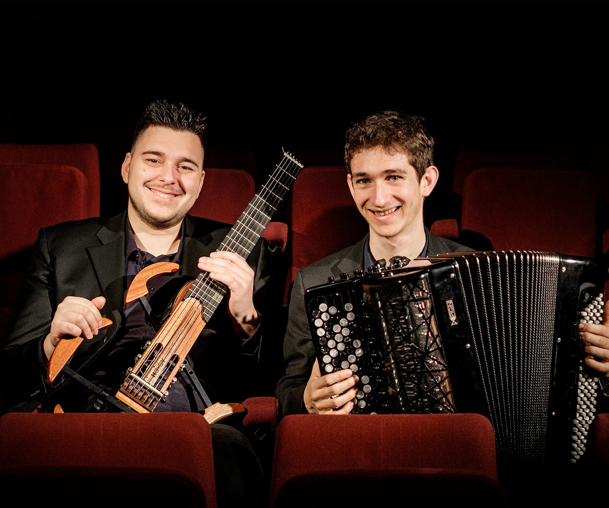 Sotiris Athanasiou avec sa guitare à la main et Julien Beautemps avec son accordéon dans les bras sont assis, souriants, dans une salle de concert.